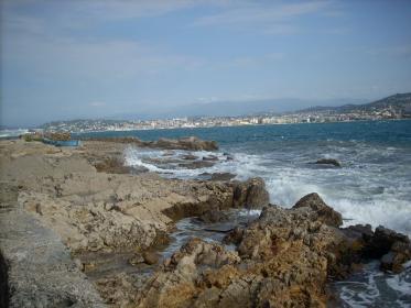 A la pointe rocheuse de l'île battue par les flots, on aperçoit la ville de Cannes
